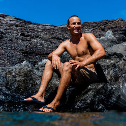 Man wearing trench blue swim shorts sitting on rocks by water.