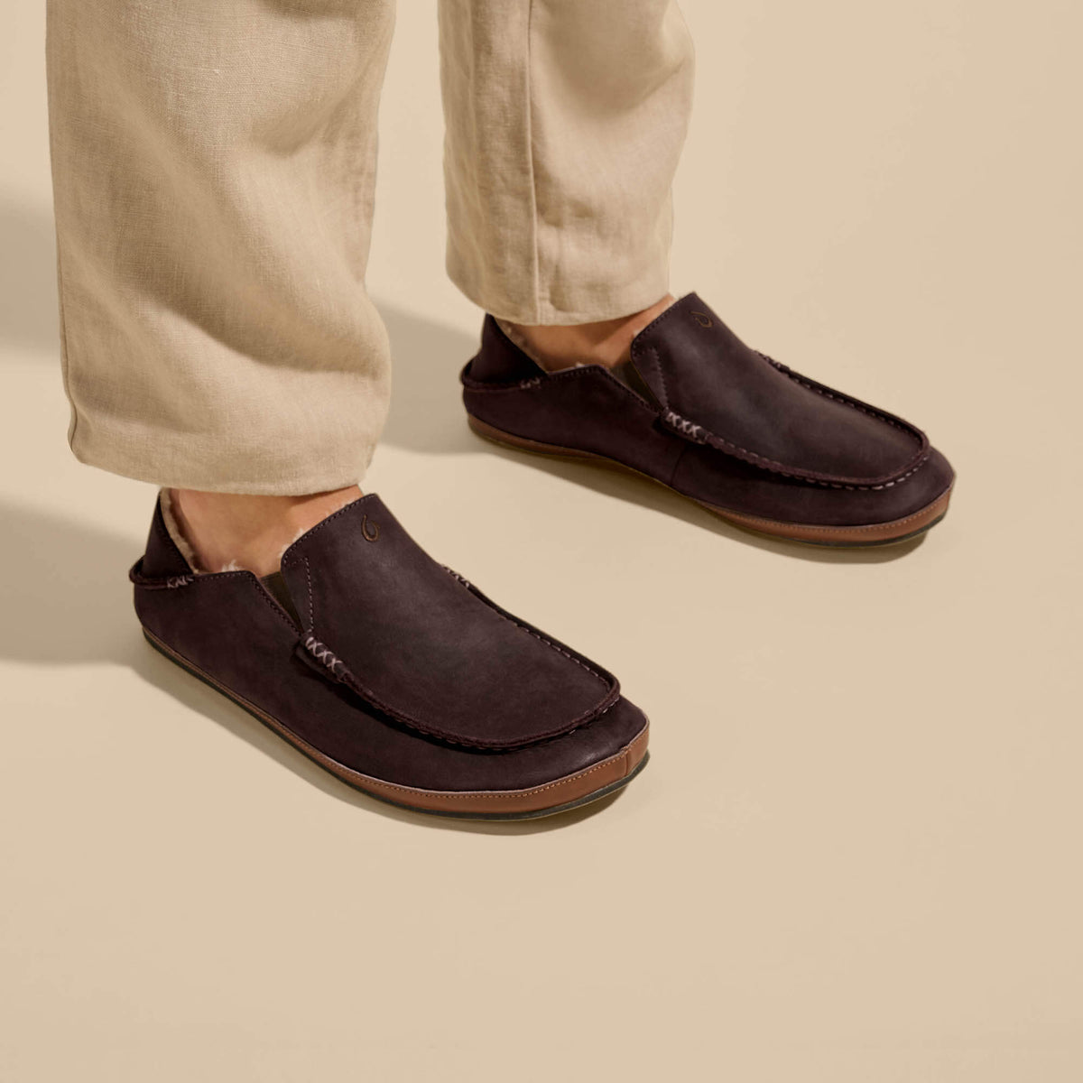Dark brown moccasin slippers with suede uppers and tan leather soles, worn with cream pants.