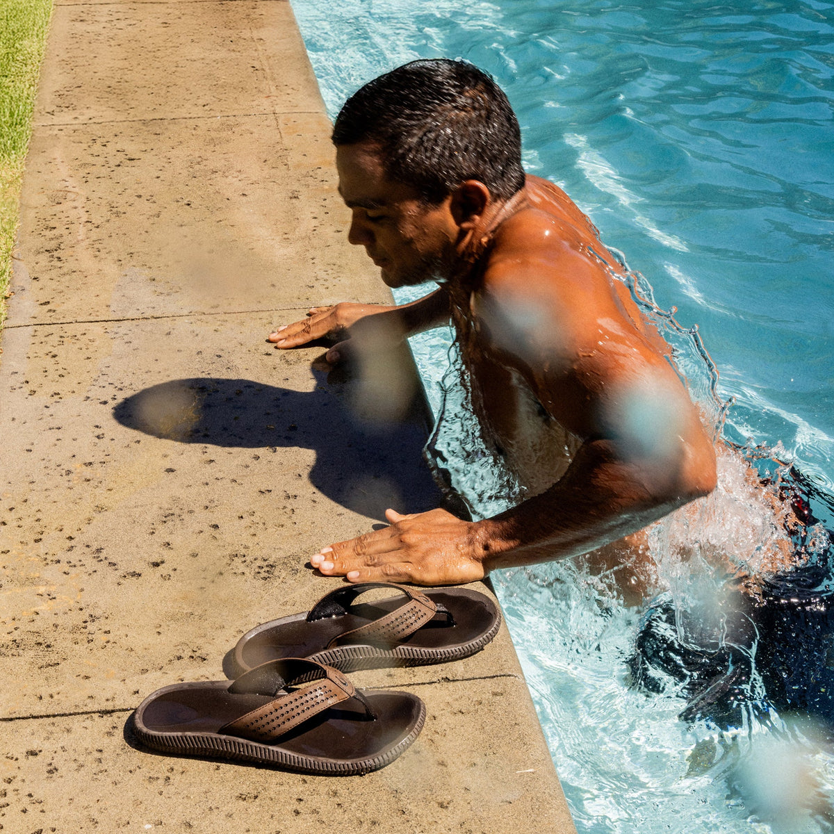 Dark Java thong sandals placed on pool deck next to person in water.