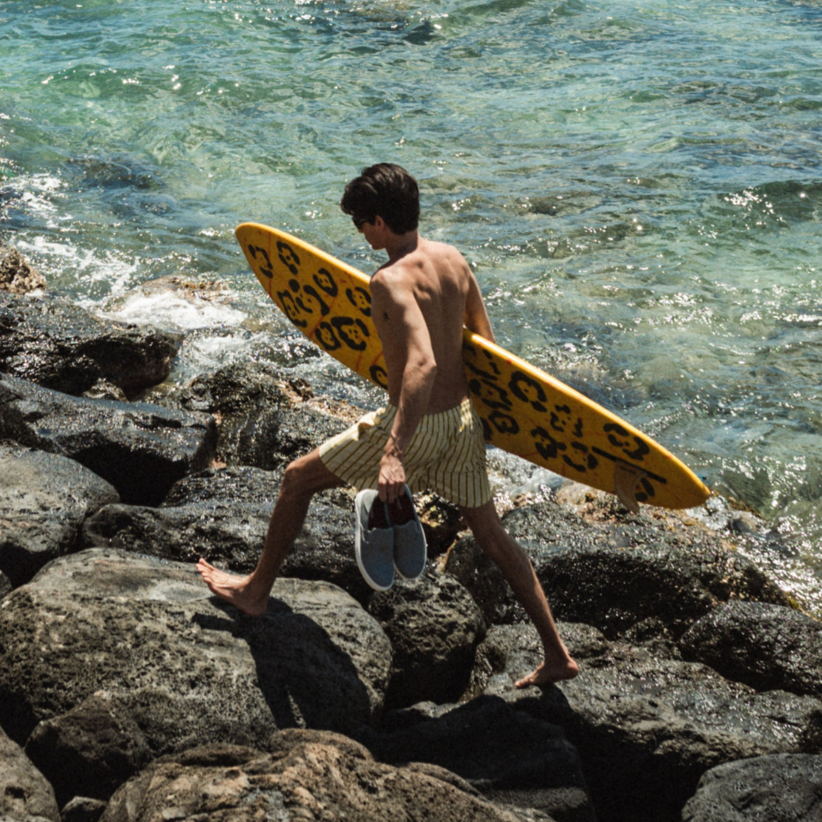 Shirtless person holding yellow surfboard with black design walking on rocky beach toward ocean.