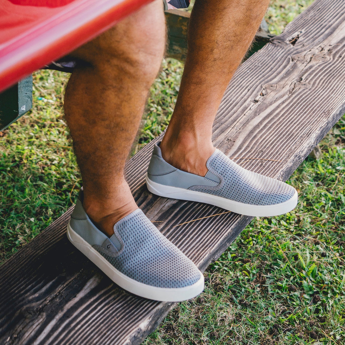 Man wearing gray perforated slip-on sneakers with white sole, standing on wooden deck near grass.