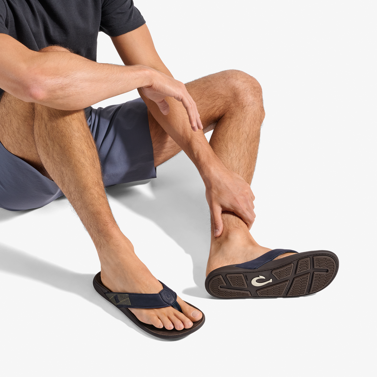 Man wearing dark shorts and navy thong sandal with dark sole, shown from waist down in seated position.