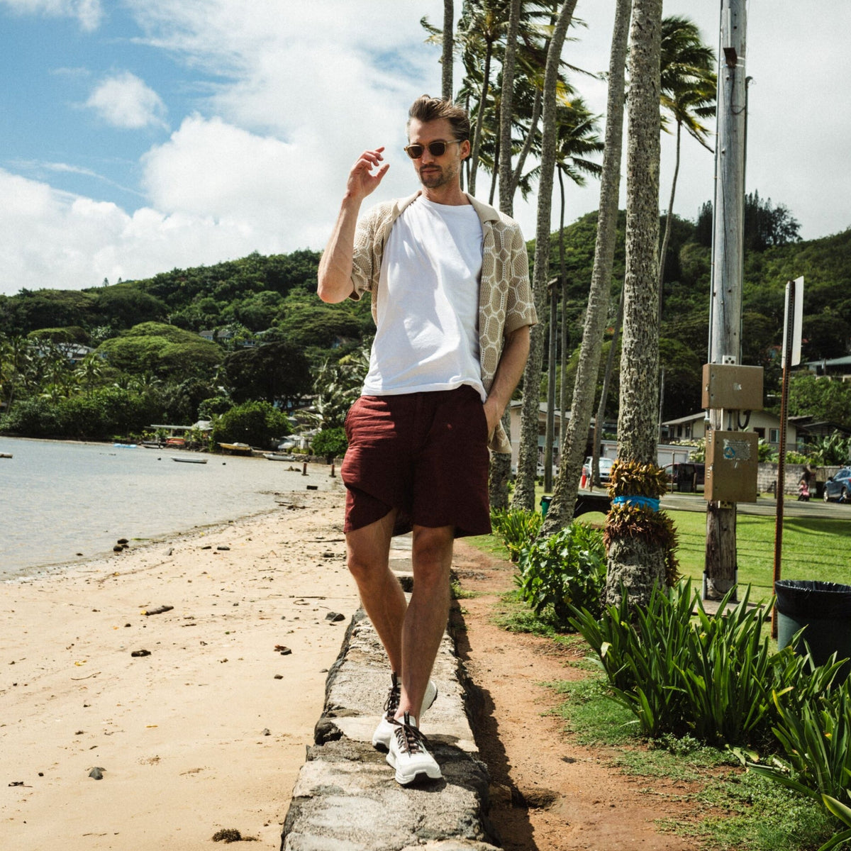 Man wearing white t-shirt, tan patterned shirt, and burgundy shorts standing on beach with palm trees and tropical landscape.