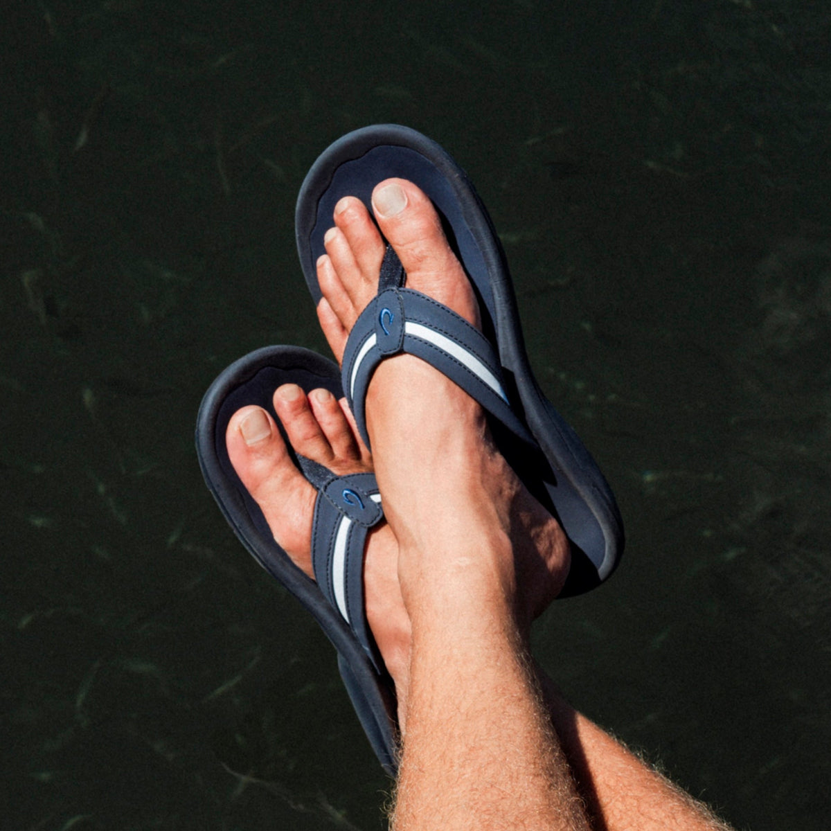 Navy thong sandals with white stripe detail on foot, worn on male feet against dark water background.