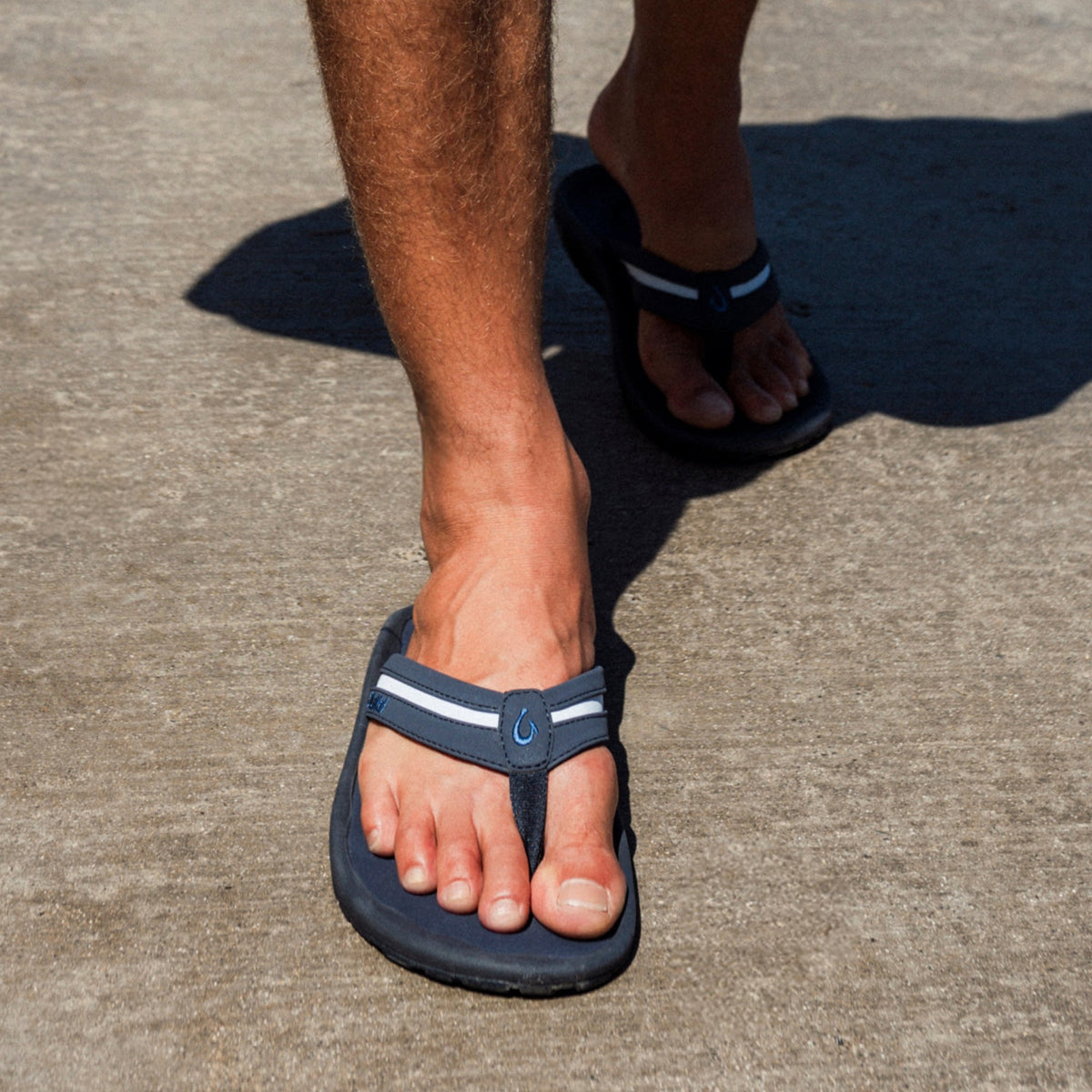 Man wearing navy thong sandal with white stripe and blue buckle detail, shown on pavement.