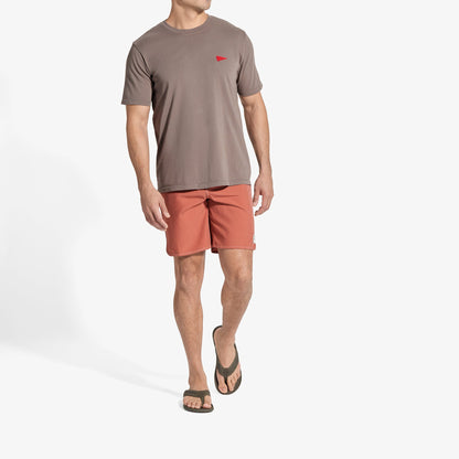 Man wearing taupe crew neck t-shirt with red flag logo and coral shorts with black sandals.