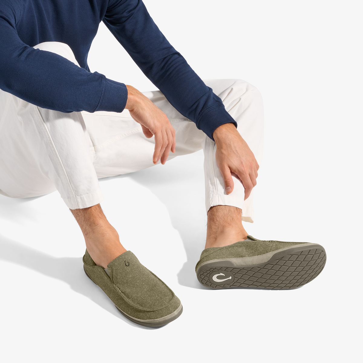 Person wearing navy long-sleeve shirt and white pants seated, displaying clay-colored Moloā Hulu slippers with textured sole.