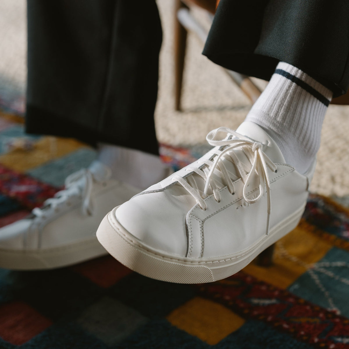 White lace-up sneaker with ribbed white socks and black pants on a patterned rug.