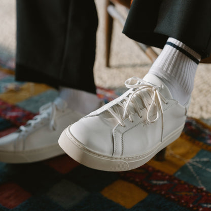 White lace-up sneaker with ribbed white socks and black pants on a patterned rug.