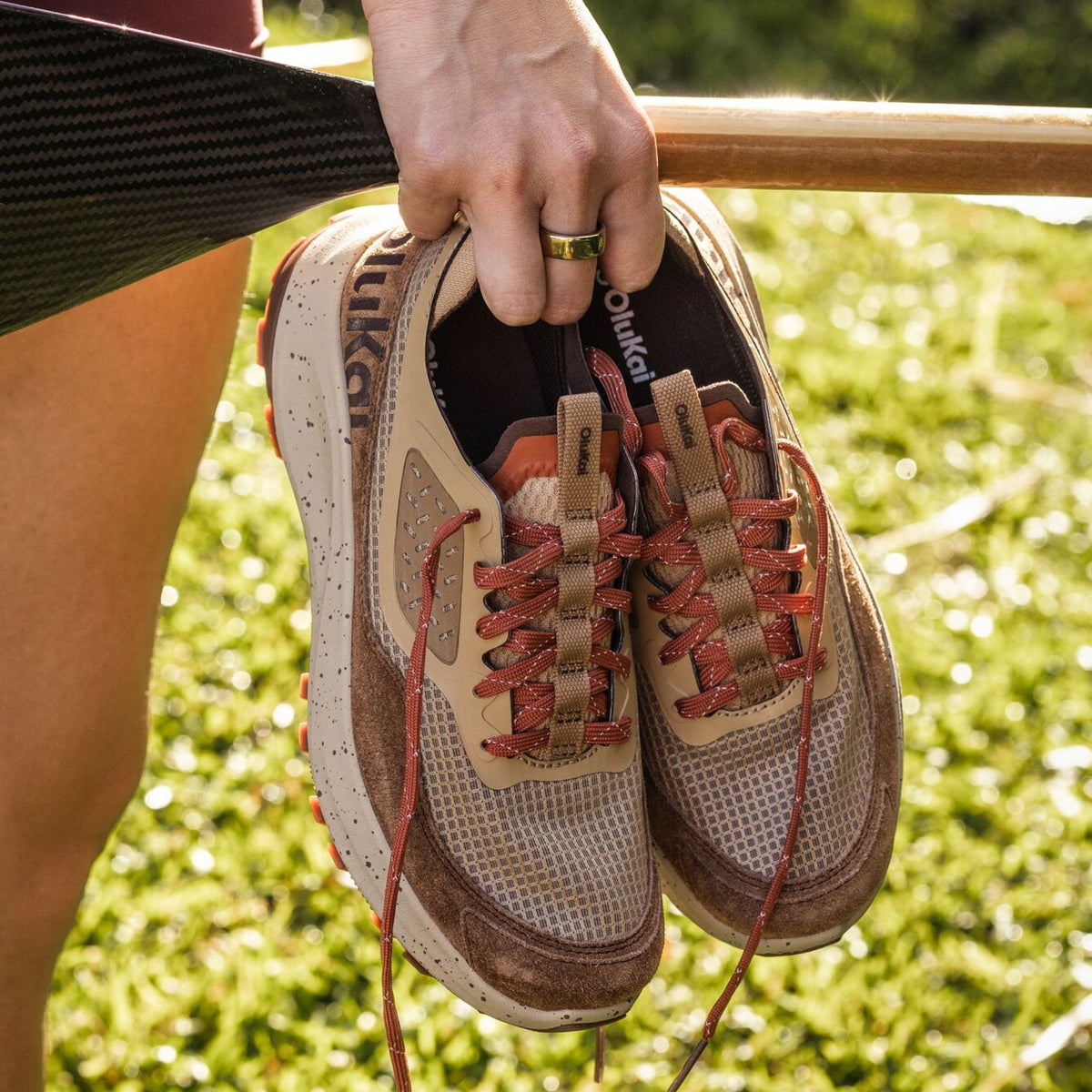 Beige and brown mesh running shoe with carob trim, red laces, and perforated details, held in hand outdoors.