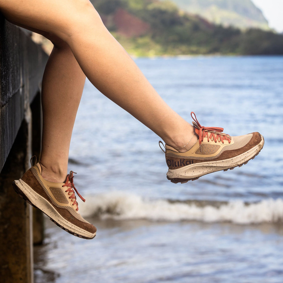 Person wearing tan and brown lace-up sneakers with red accents, dangling feet over water.
