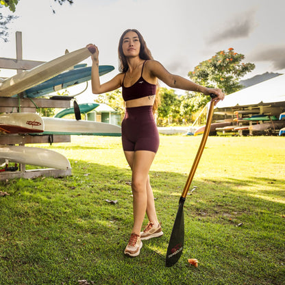 Woman wearing carob-colored sports bra and sandshell shorts holding a paddle outdoors.