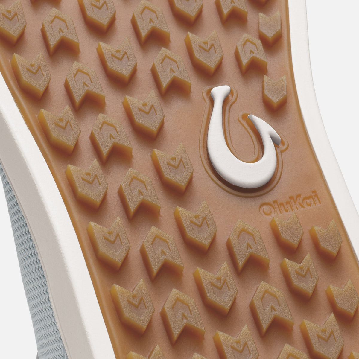 Tan rubber sole with chevron tread pattern, white Olukai heel logo, and grey mesh upper visible at edge.